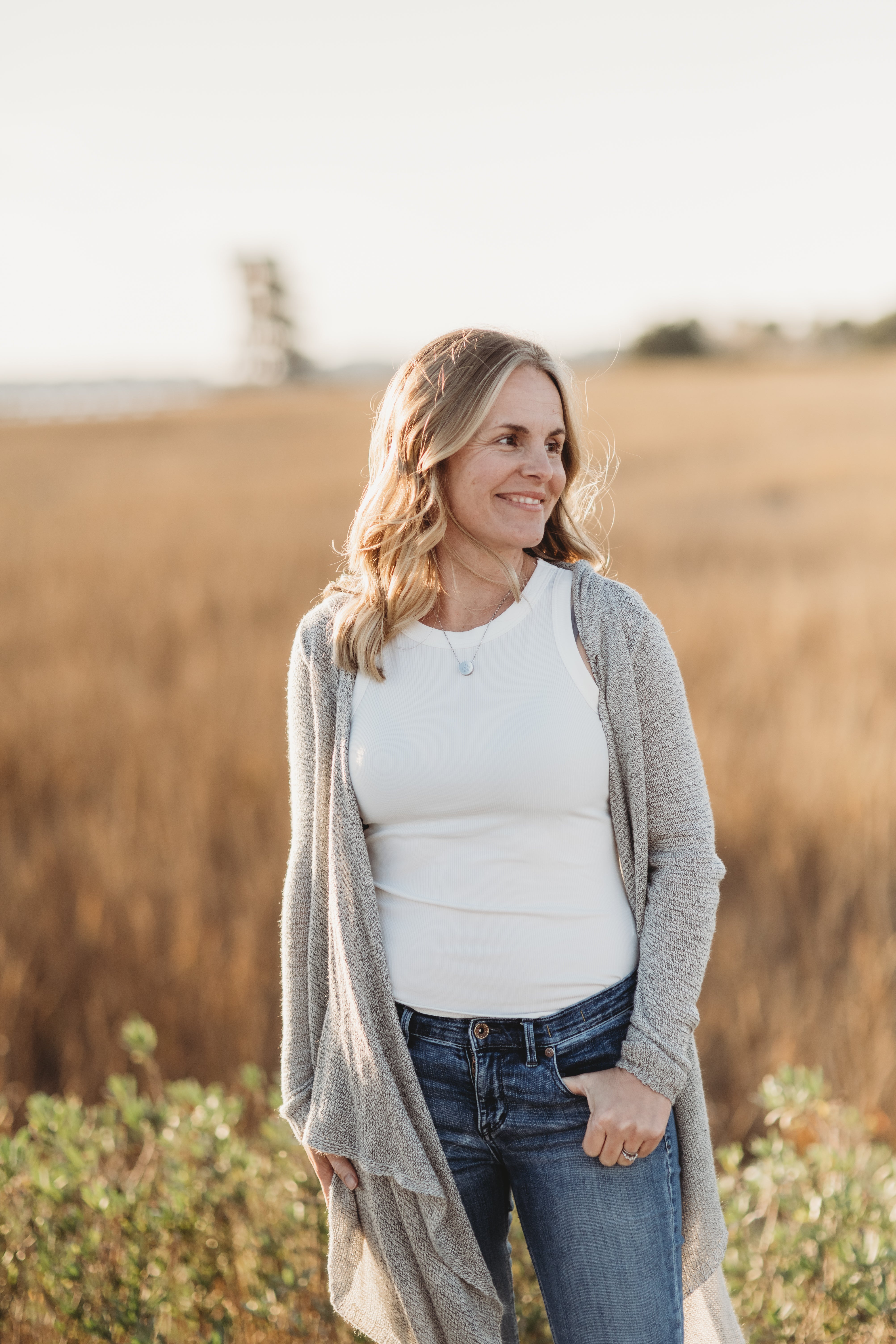 A woman standing in a field wearing a long cardigan, a white shirt, and jeans. - Lactation Consulting Services
