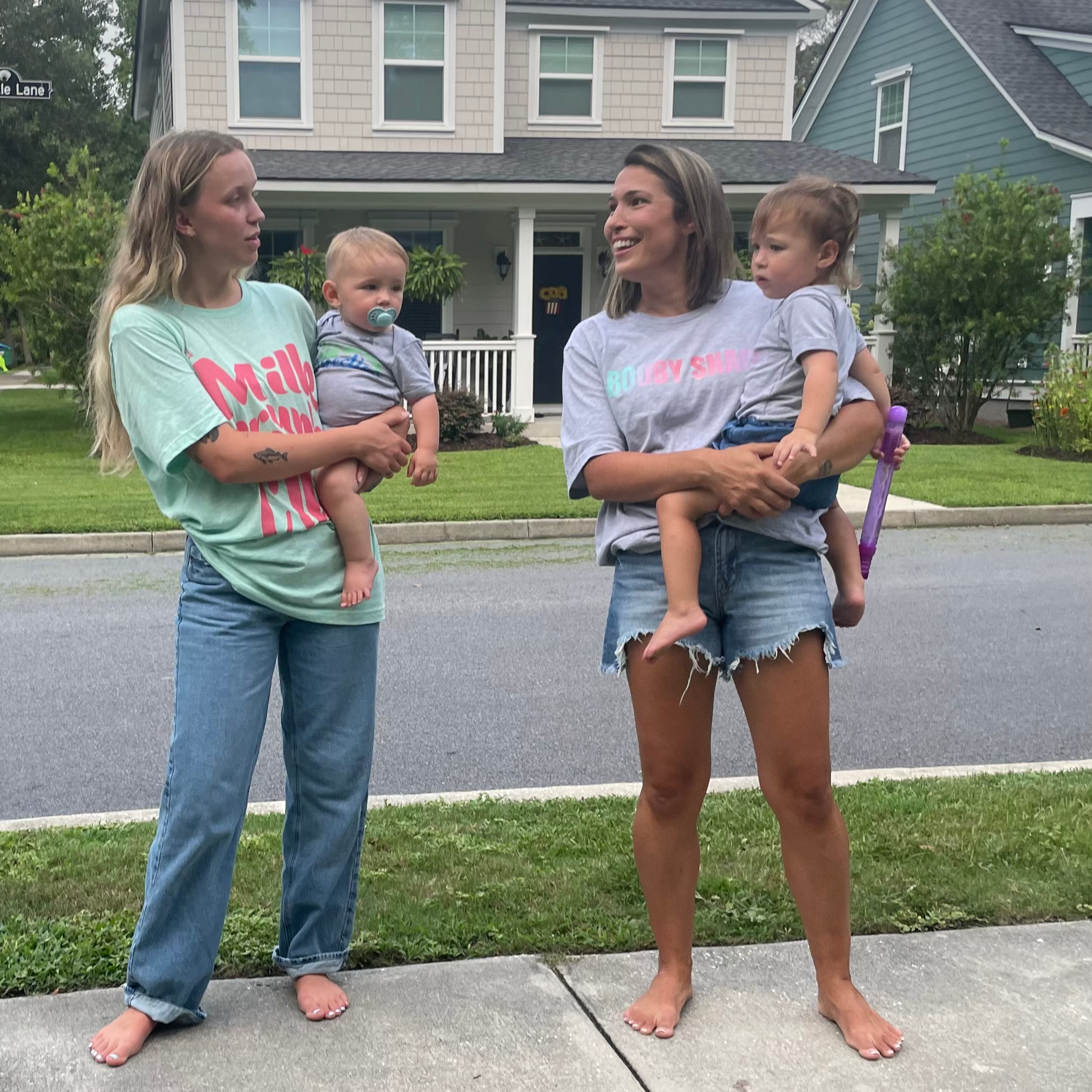 Two women are standing on a sidewalk holding babies in front of a house.