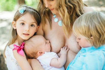 Woman breastfeeding a baby with two children looking on outdoors.