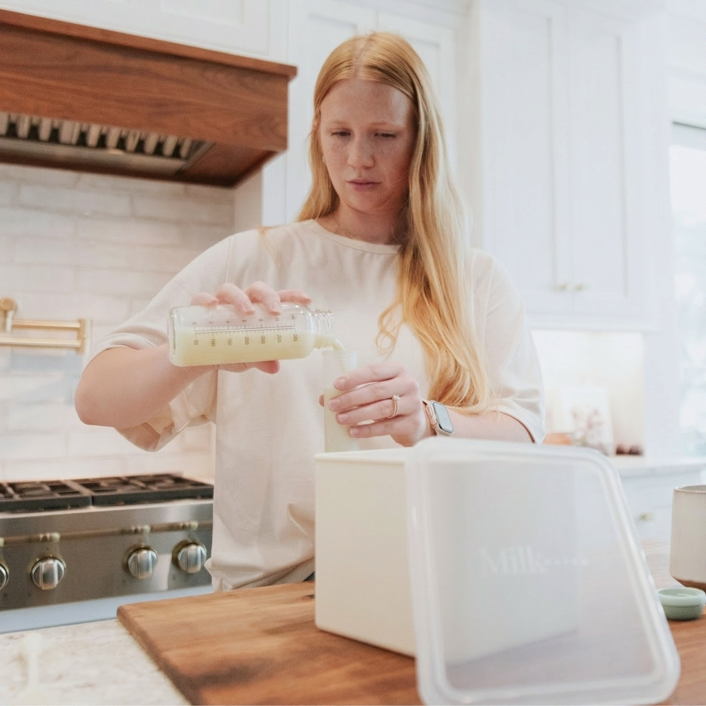 A woman in a cream-colored top stands in a bright kitchen, pouring milk from a glass measuring container into a silicone milk storage container with a clear lid. A wooden cutting board and stainless steel stove are visible on the counter.